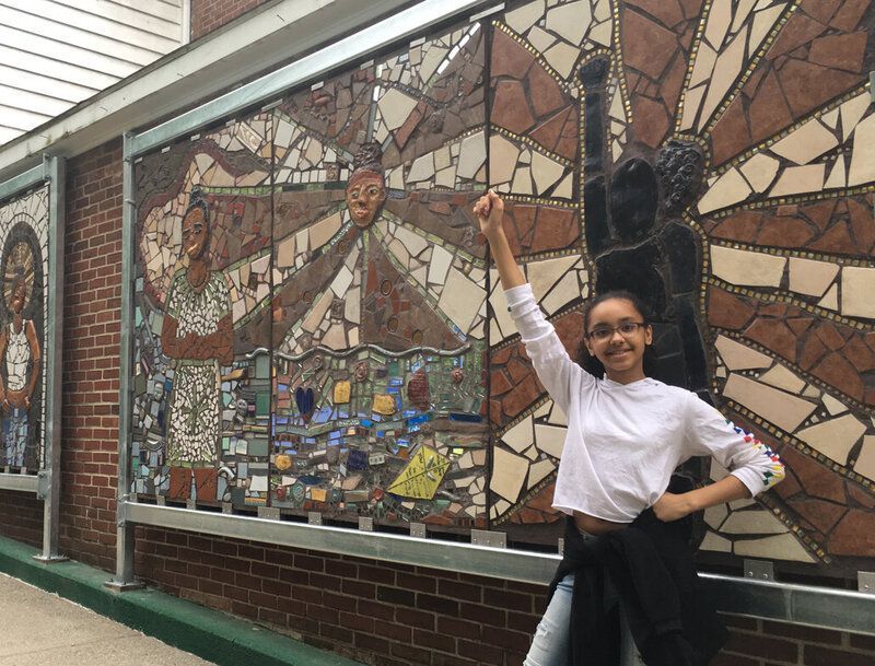 A young girl is standing in front of a mosaic wall.