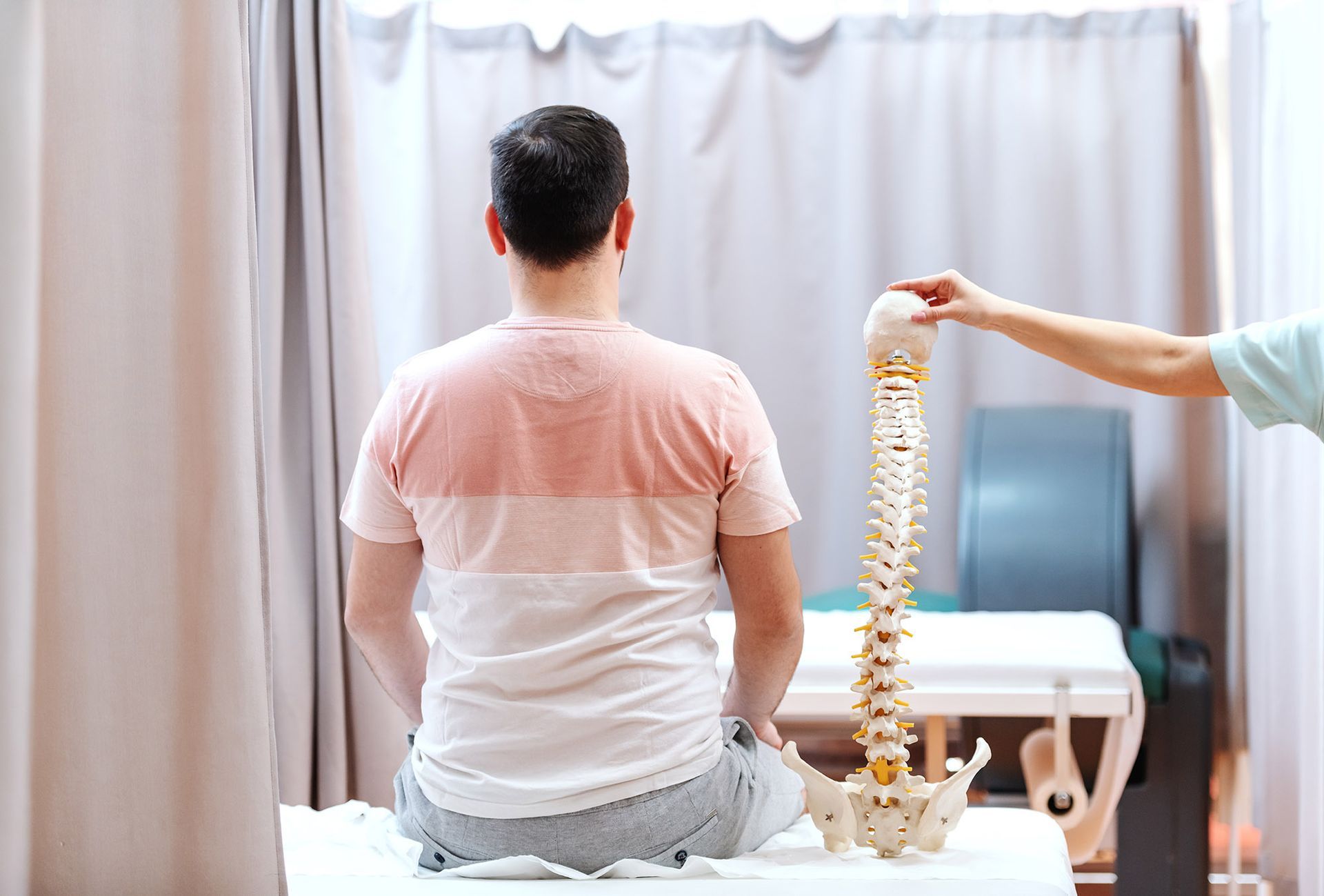 Person seated while another holds a spine model, in a clinic setting.
