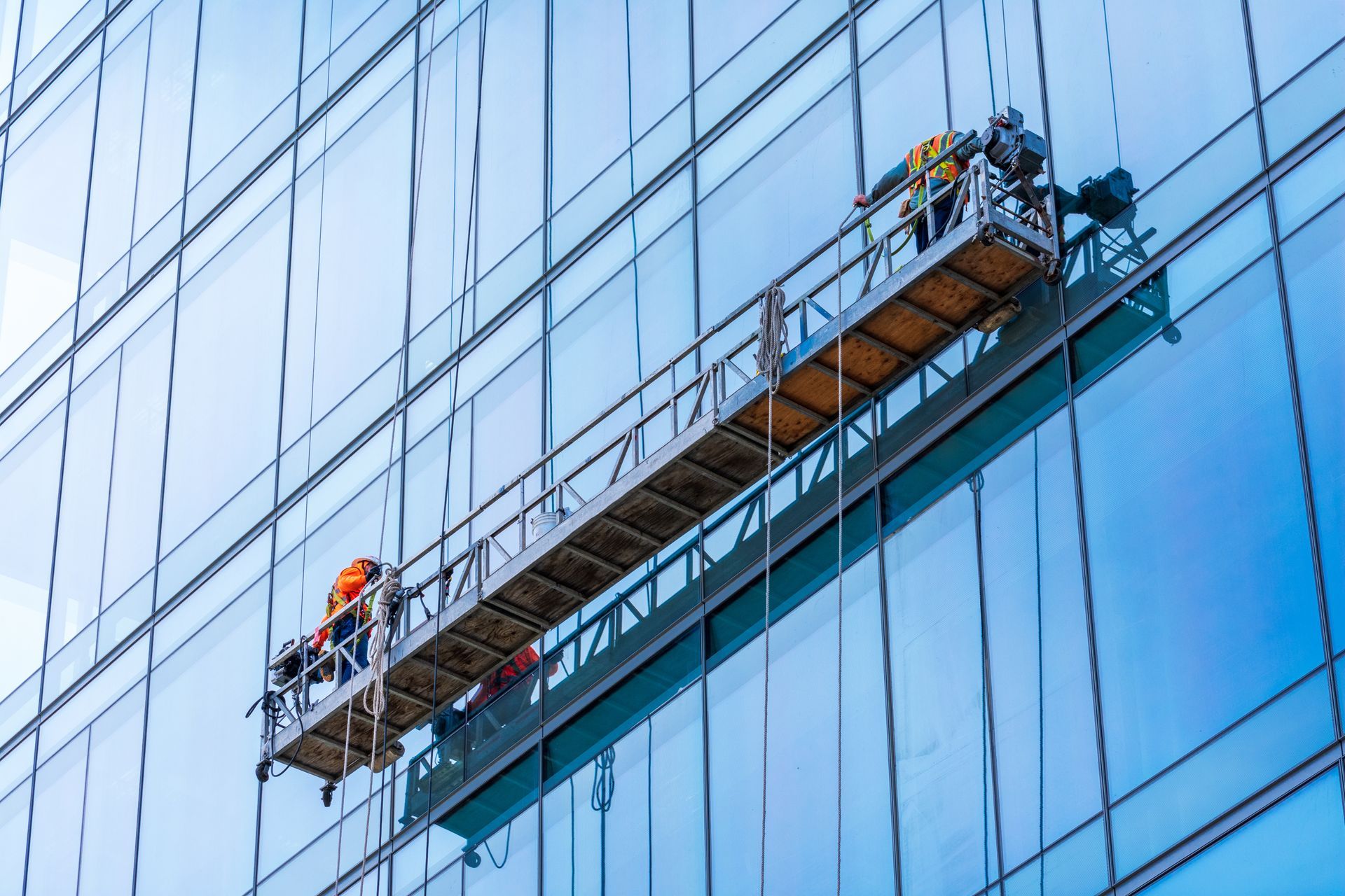 Workers on a suspended platform cleaning a large glass building facade.