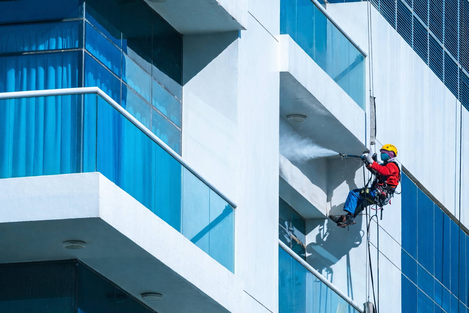 A window washer in a harness sprays water on a tall building, blue glass and white walls.