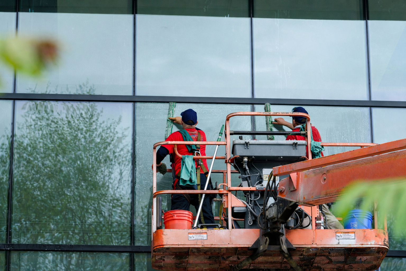 Two window cleaners in red uniforms work from a raised orange platform to clean a large glass building facade.