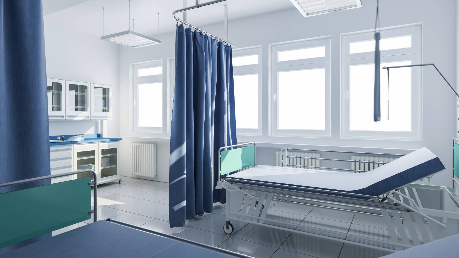Hospital room with beds, blue curtains, and medical equipment. Bright room with windows.