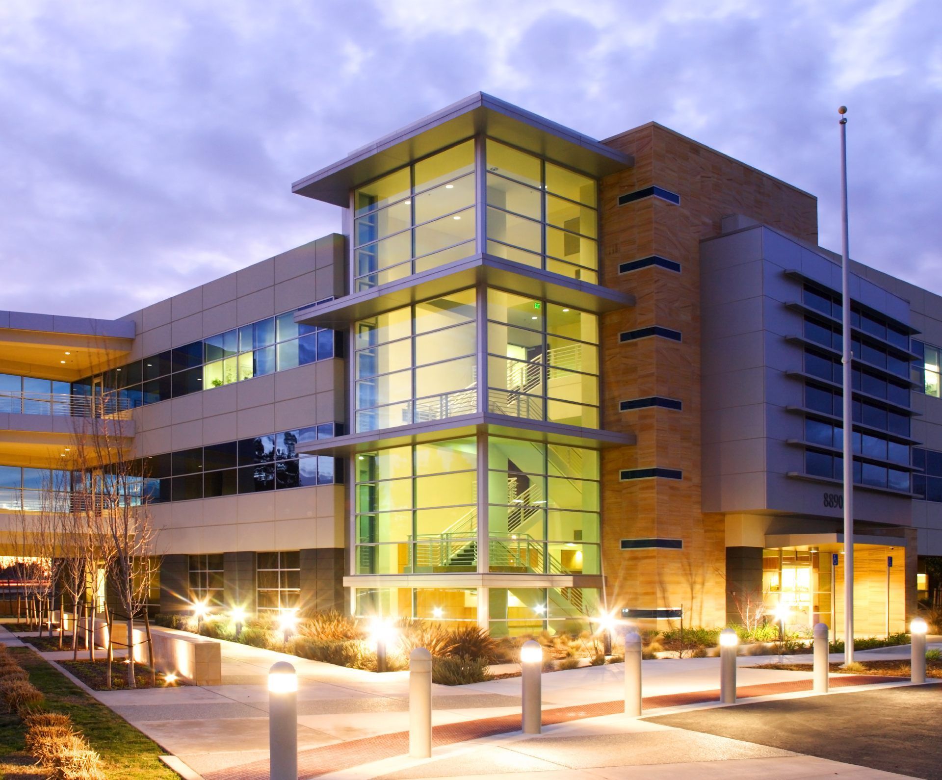 A modern three-story office building with a glass-enclosed staircase, glowing internal lights, and evening landscaping.