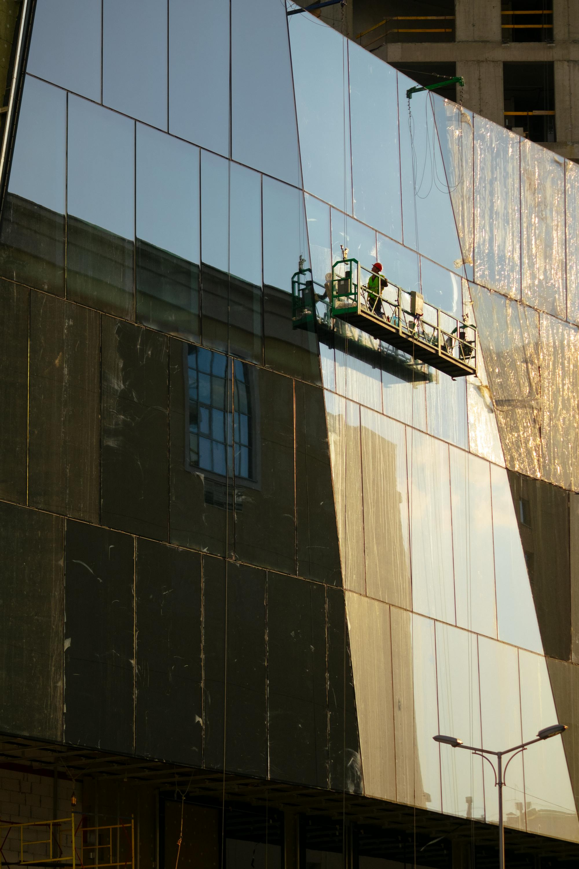 Workers on a suspended platform clean the glass facade of a modern building, which reflects the surrounding architecture.