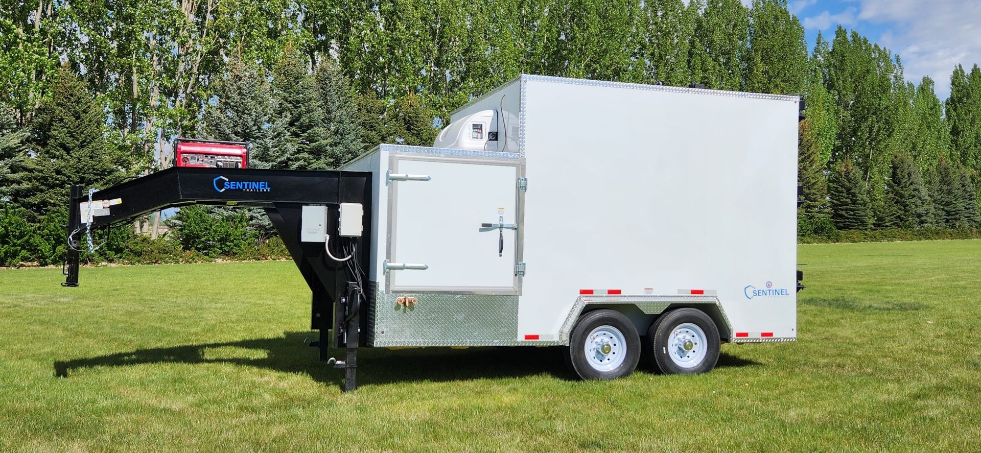 Black refrigerated trailer with white enclosed cargo box parked on grass beside trees