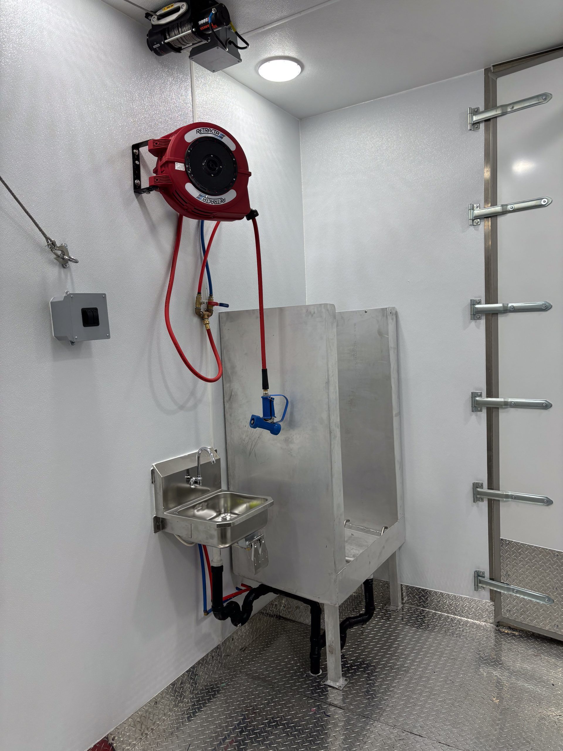 Small industrial stainless-steel washing station in a white room, with red hose reel and wall-mounted fixtures.