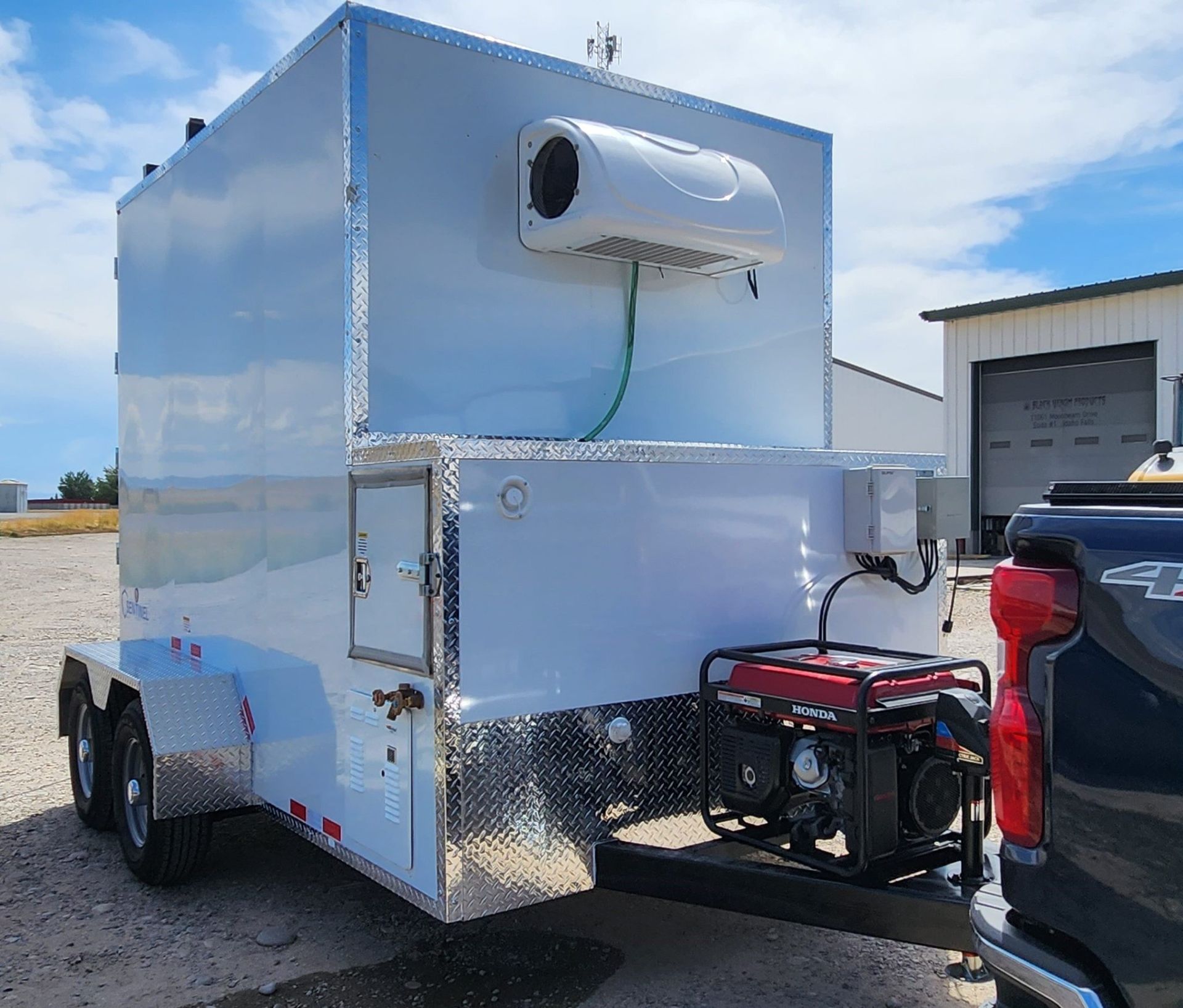 Silver enclosed trailer with a rooftop HVAC unit, parked beside a pickup truck in a lot.
