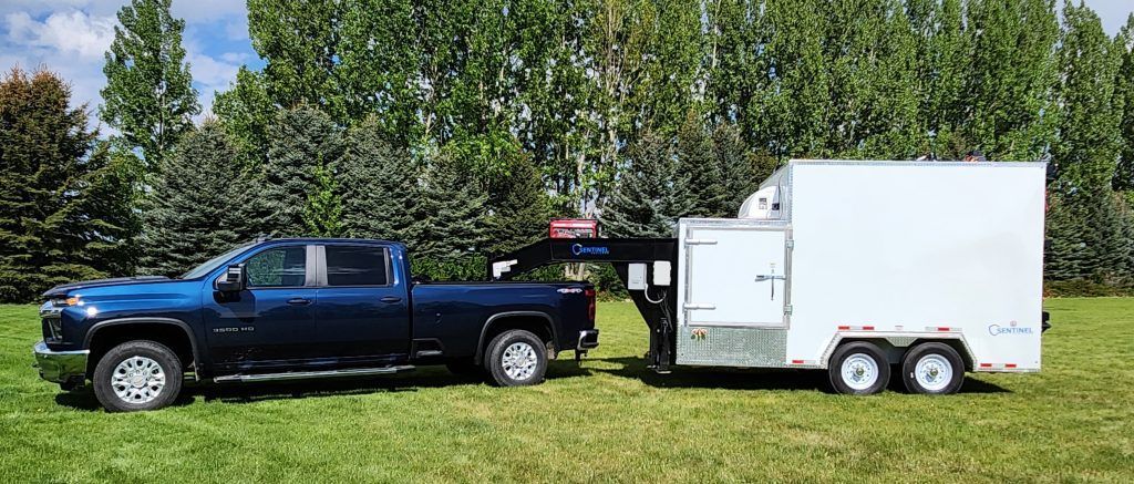 Blue pickup truck towing a butcher trailer on a grassy field with trees in the background