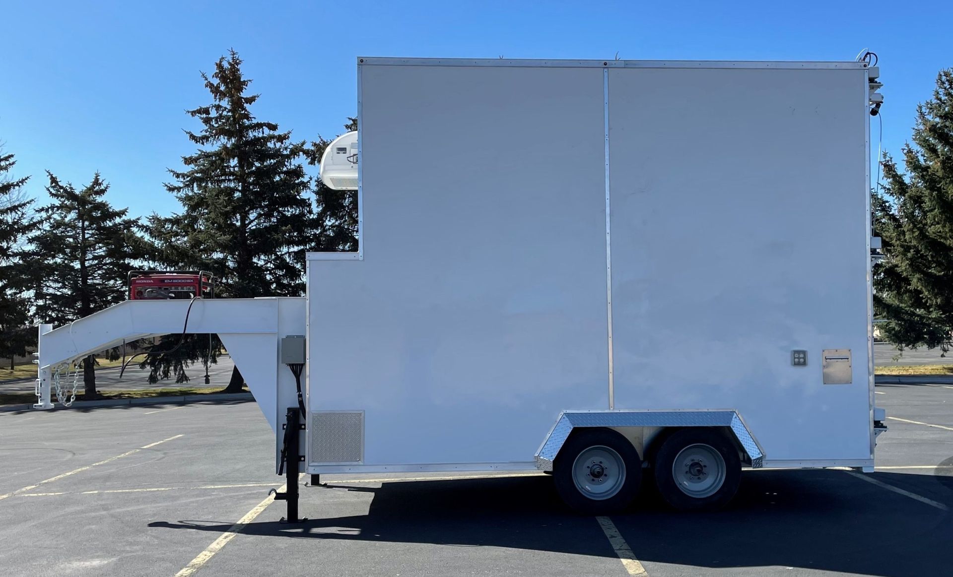 White box trailer with a side awning and twin axles parked in a sunny lot, trees in the background