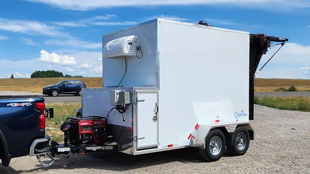 White butcher trailer with awning and generator parked on gravel beside a black pickup under a blue sky