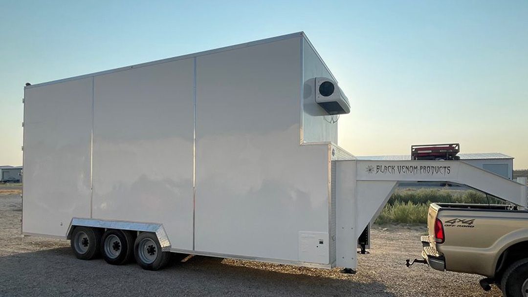 White enclosed butcher trailer with rooftop vent parked on gravel beside a pickup truck at sunset.
