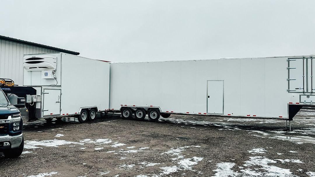 Snowy parking lot with large white refrigerated semi-trailers and a truck under an overcast sky