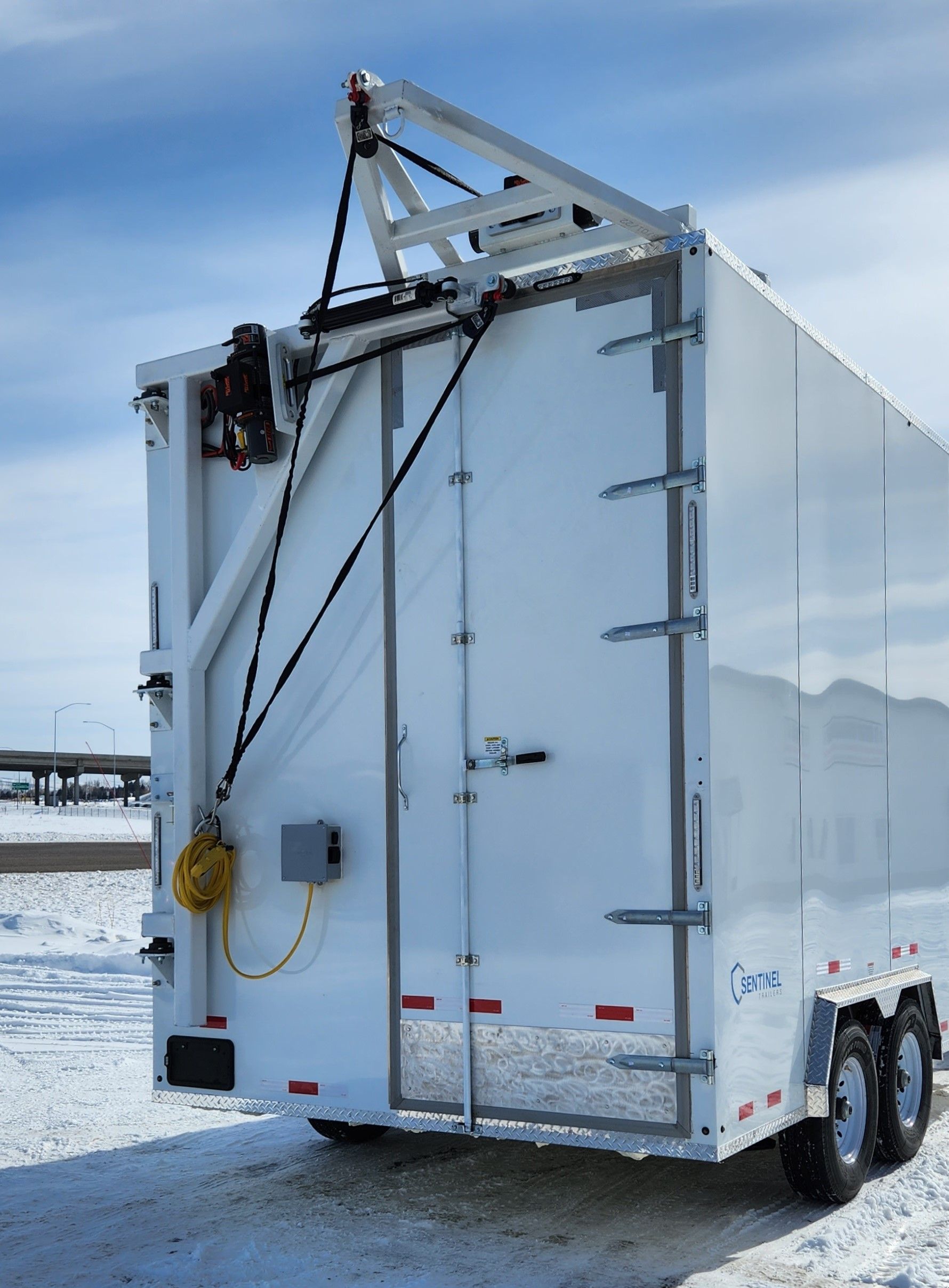 White utility trailer on snowy ground, rear view with ladder and cables under a blue sky