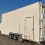 Beige mobile trailer with a side door and three axles, parked outdoors under a blue sky