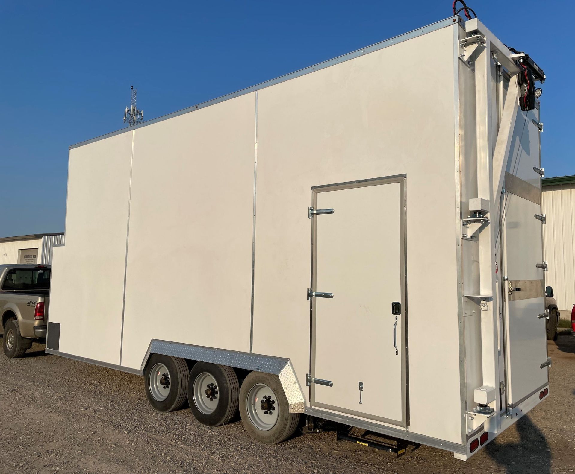 White enclosed butcher trailer with triple axles and side door parked on gravel under a blue sky.