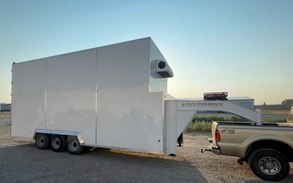 White box trailer for butchers with a side vent parked on gravel beside a pickup truck under a clear sky.