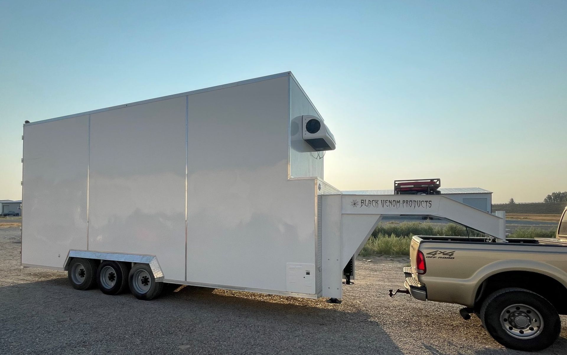 White enclosed trailer hitched to a pickup truck in a gravel lot at sunset.