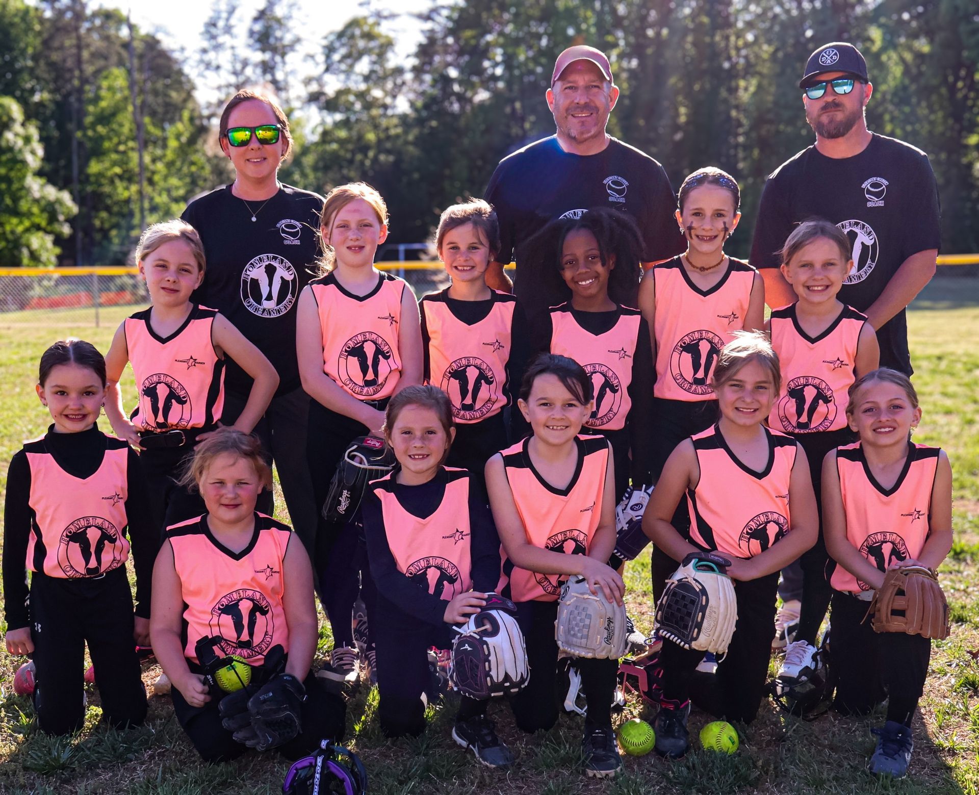 Youth sports team posing outdoors in pink jerseys with coaches and trophies in front of trees.
