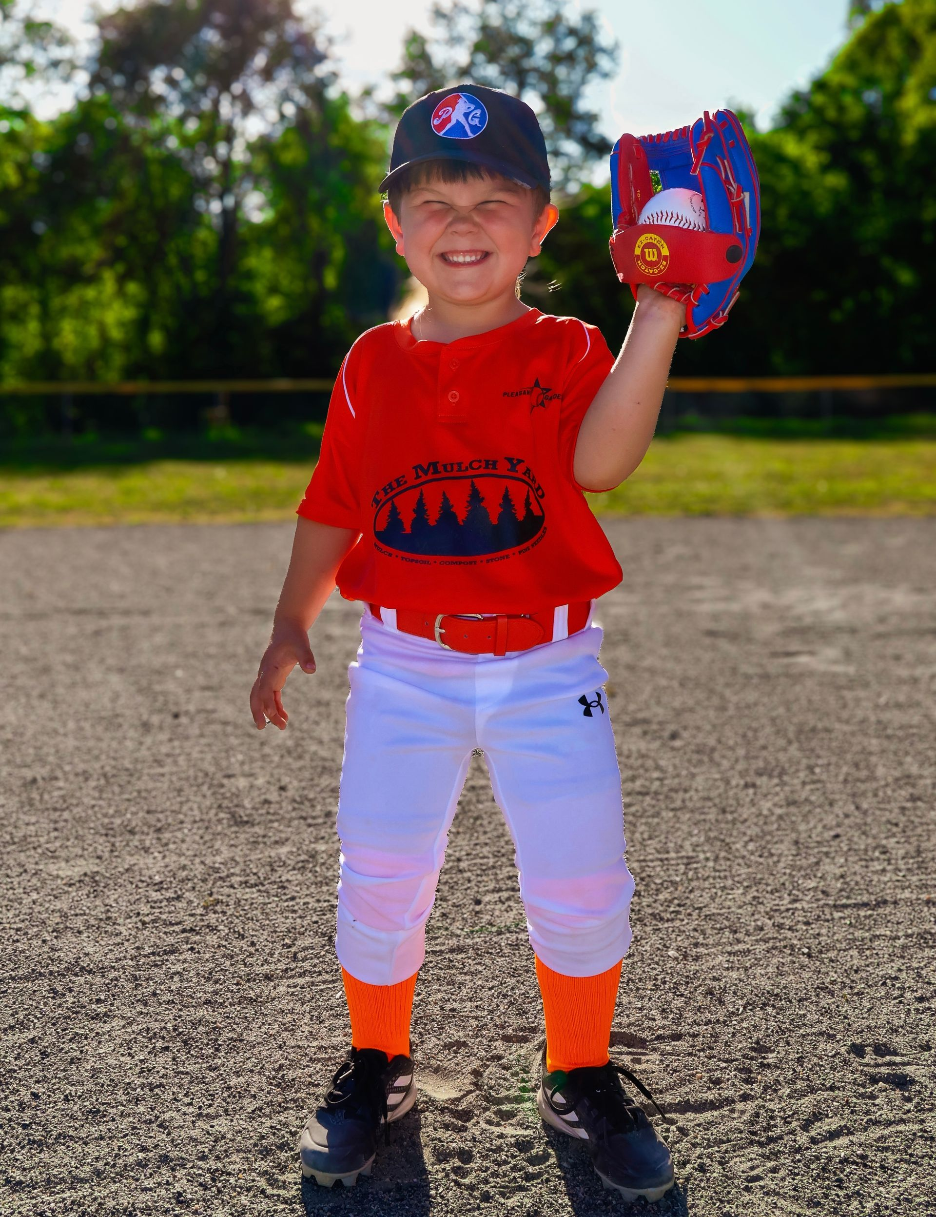 Child in red shirt and white pants smiling on a gravel field, holding a colorful toy glove.