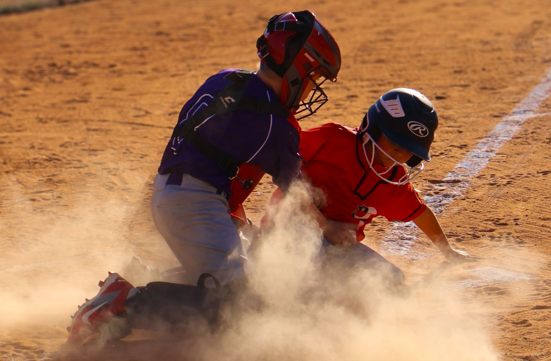 Baseball players collide at home plate, kicking up dust during a close play.
