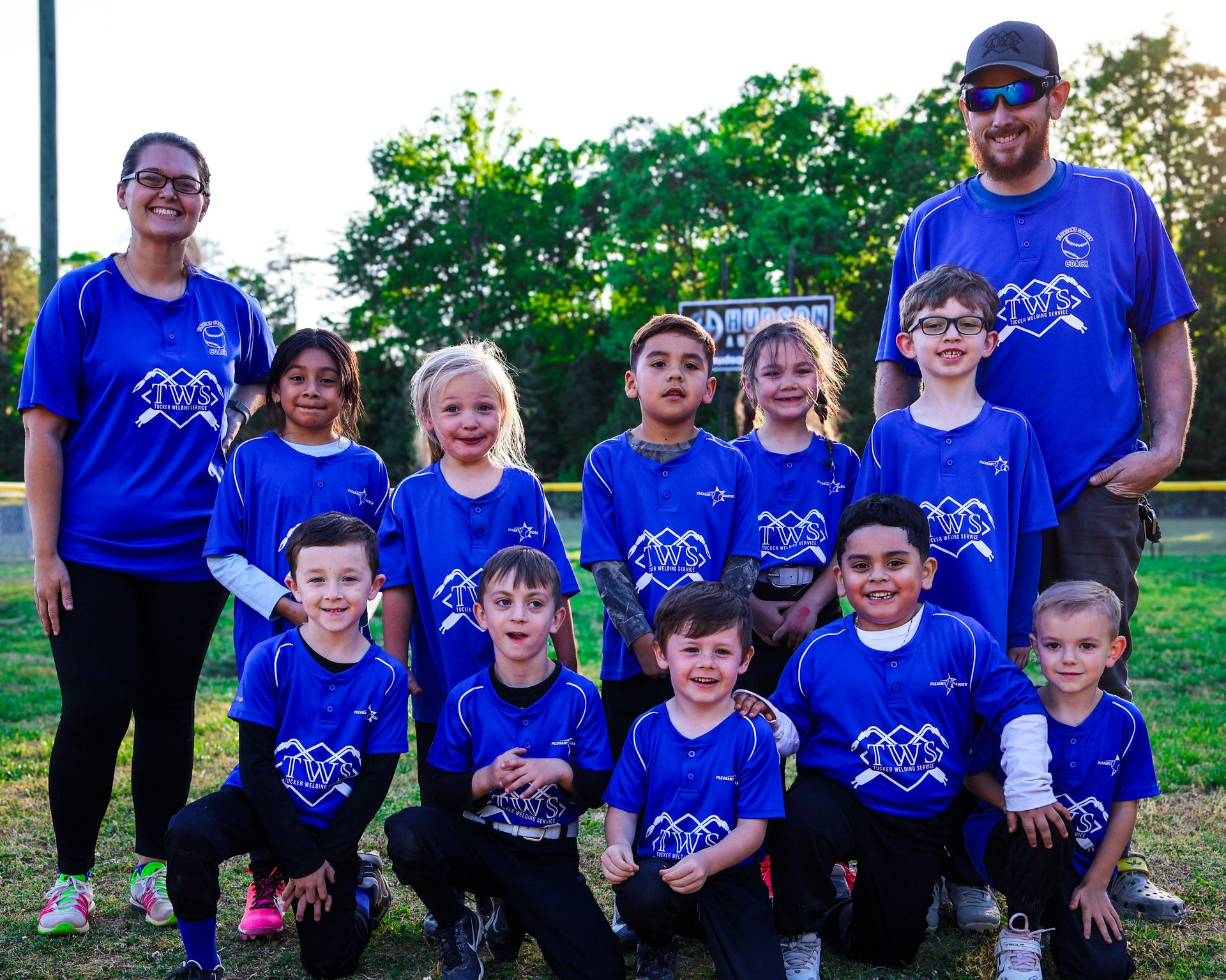 Group of children and two adults in matching blue shirts posing outdoors in a grassy field.
