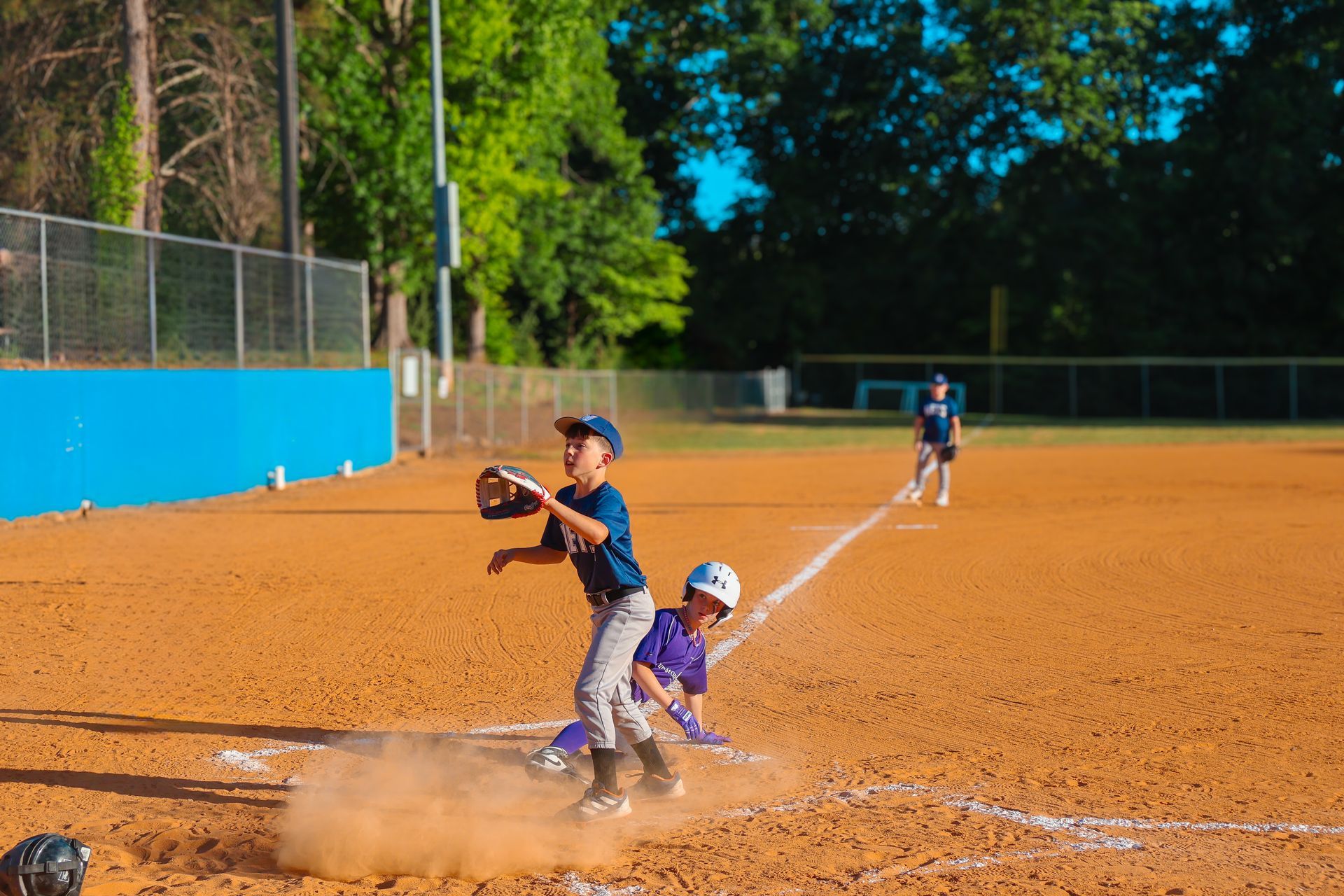 Youth baseball play at a dirt field, with a runner sliding into home as a catcher reaches for the ball.