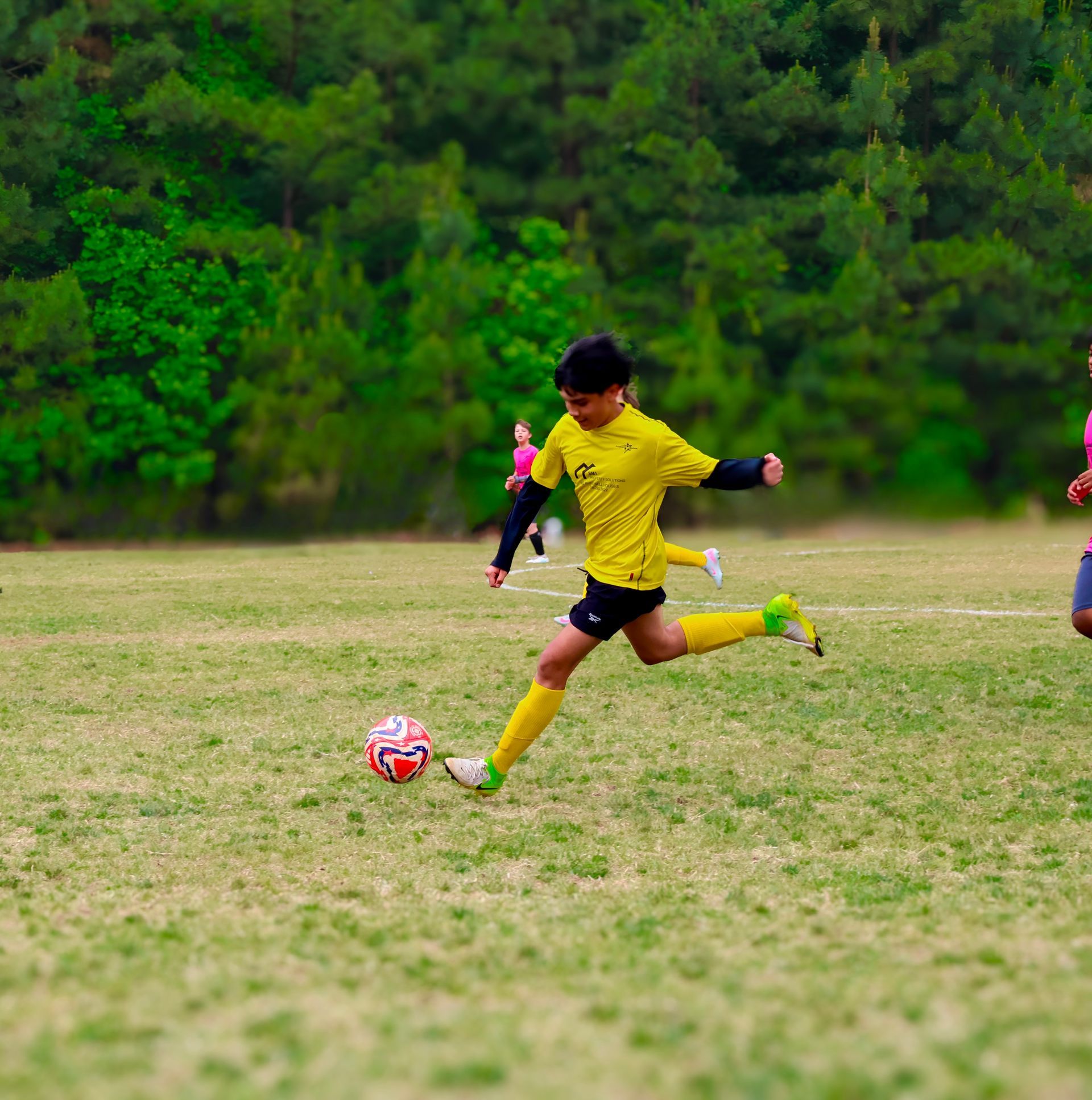 Soccer player in yellow jersey kicking a red ball on a grassy field with trees in the background