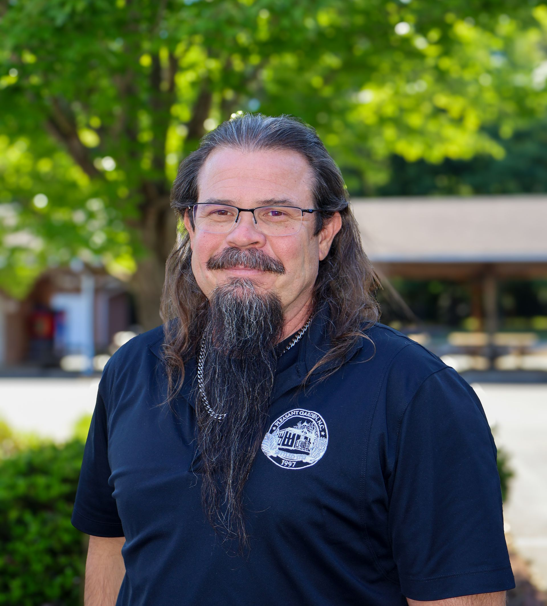 Man with glasses and long beard in a navy shirt standing outdoors in front of trees and a building
