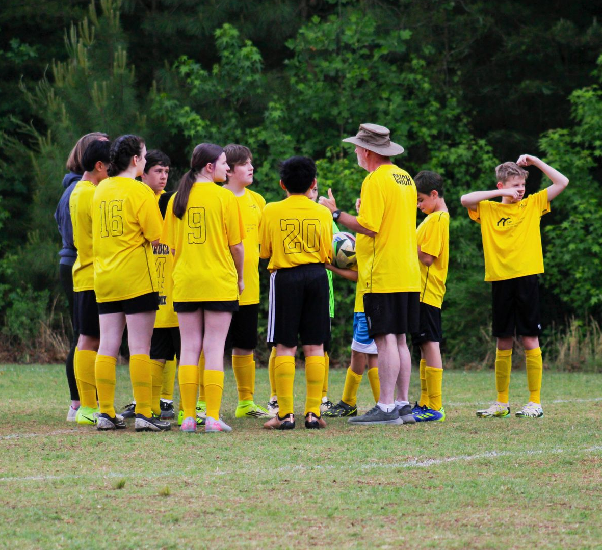 Youth soccer team in yellow jerseys huddles on a grassy field with a coach nearby.