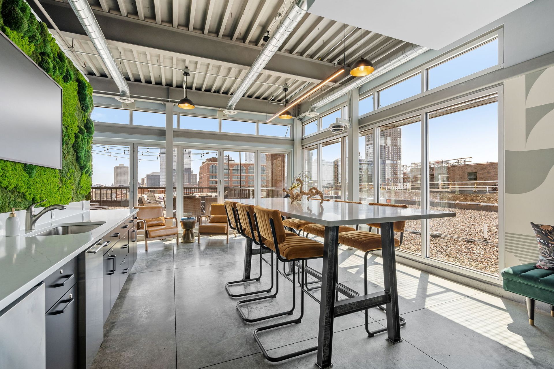 Modern indoor lounge with a long white table, tall brown stools, a green moss wall, and floor-to-ceiling windows.