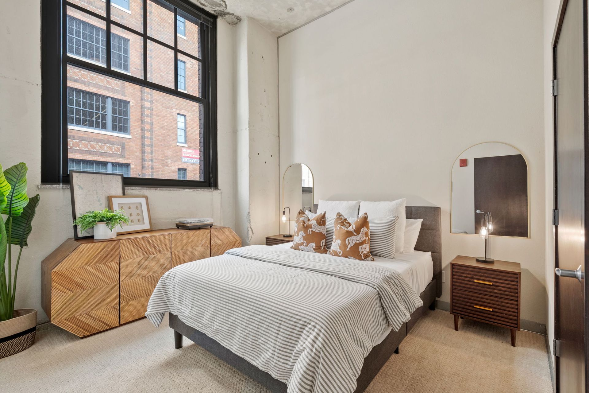 A sunlit bedroom features a bed with striped bedding, a wooden sideboard, and a small nightstand by a large window.