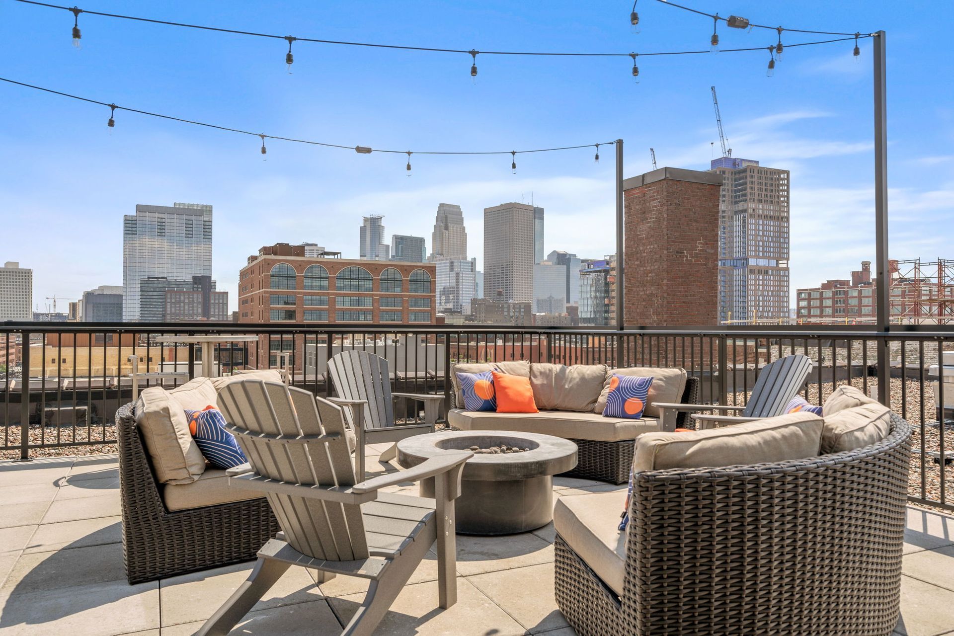 A rooftop patio with outdoor lounge seating, a fire pit, and string lights, set against a backdrop of a city skyline.