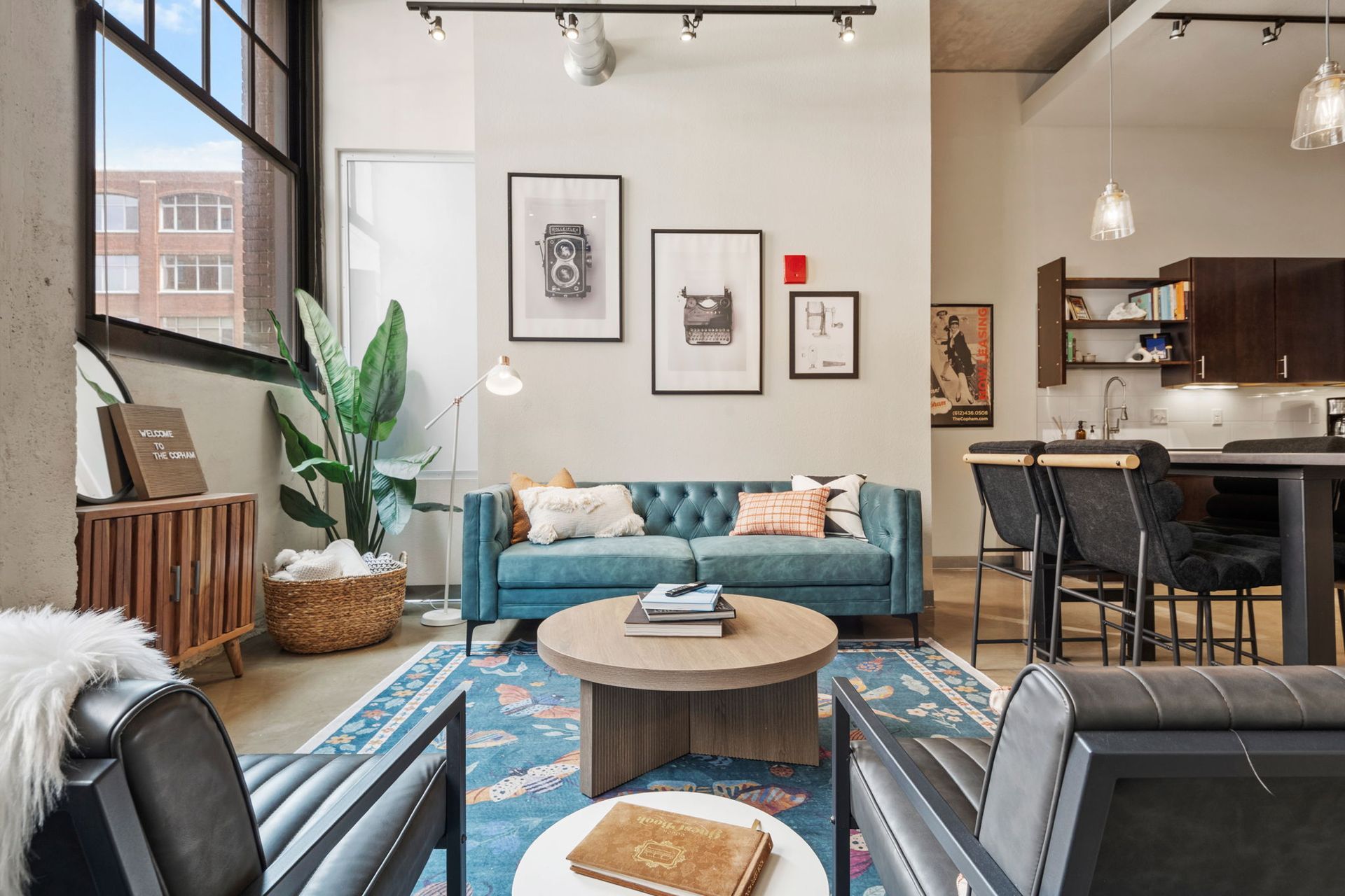 An open-plan apartment living area with a blue sectional sofa, a checkerboard rug, and a modern kitchen with gray cabinets.