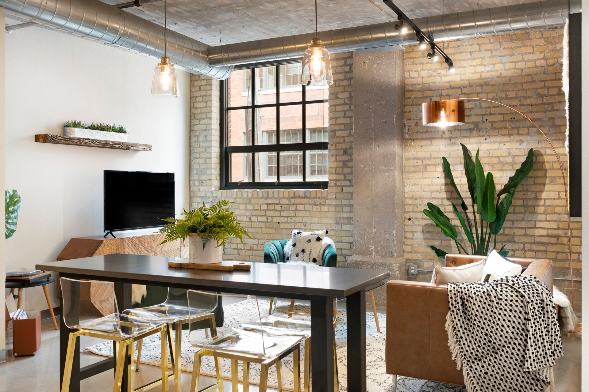 A modern apartment living room with brick walls, a dining table with gold chairs, a tan couch, and a wall-mounted TV.