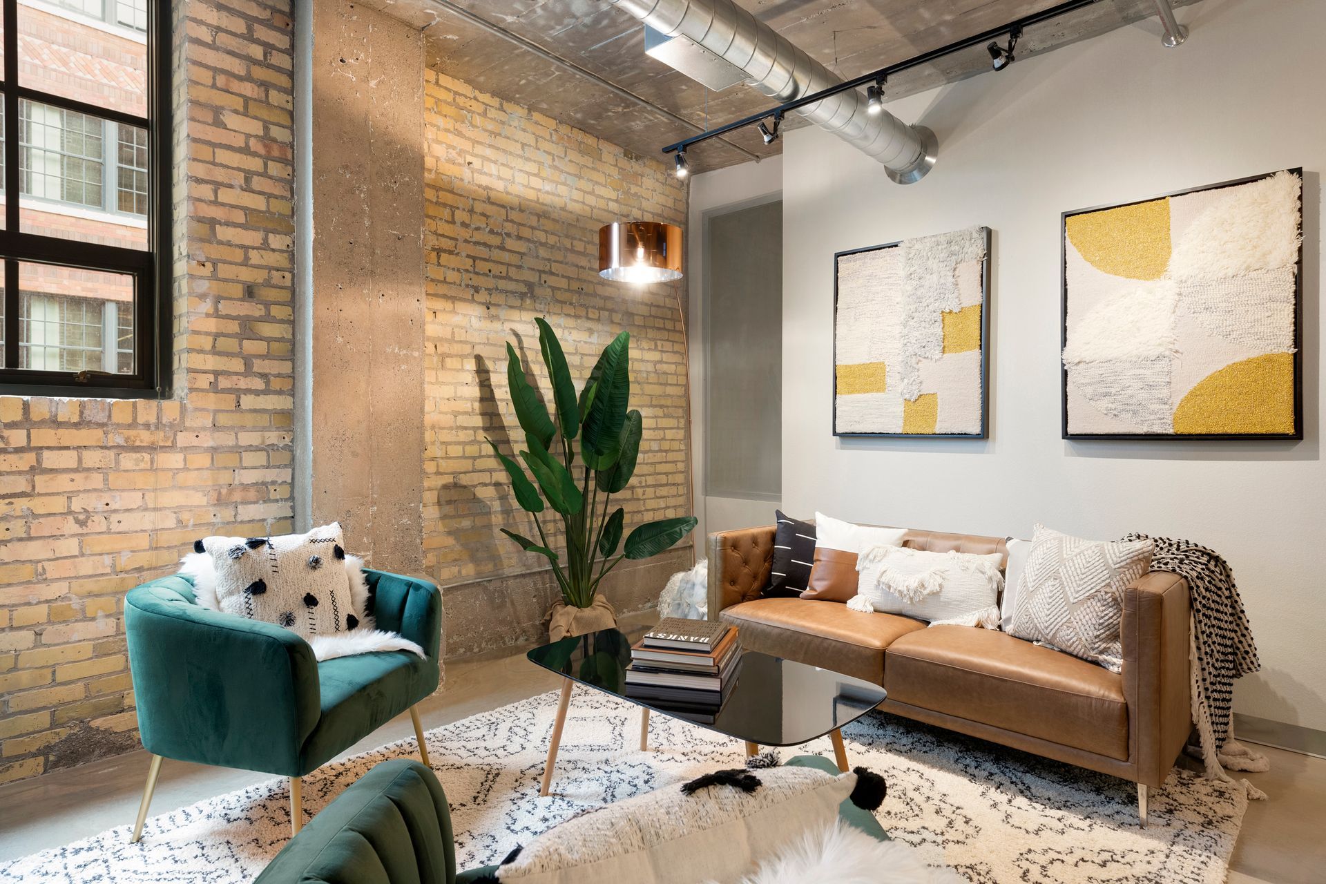 A modern lounge with a brown sofa, a green velvet armchair, a glass coffee table, and abstract art on a brick wall.