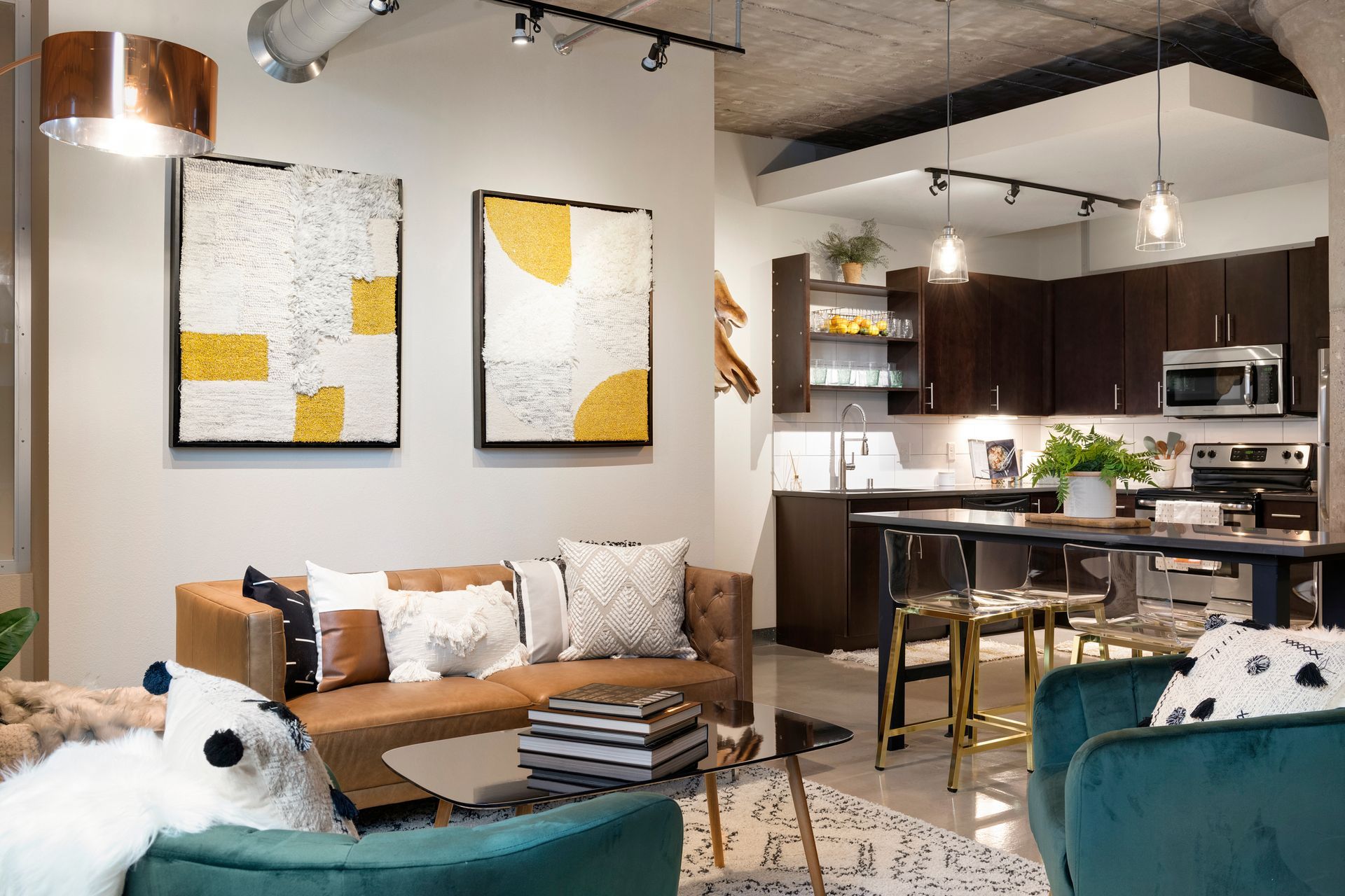 A modern living room with a brown sofa, teal chairs, abstract art, and an open kitchen with dark cabinets.