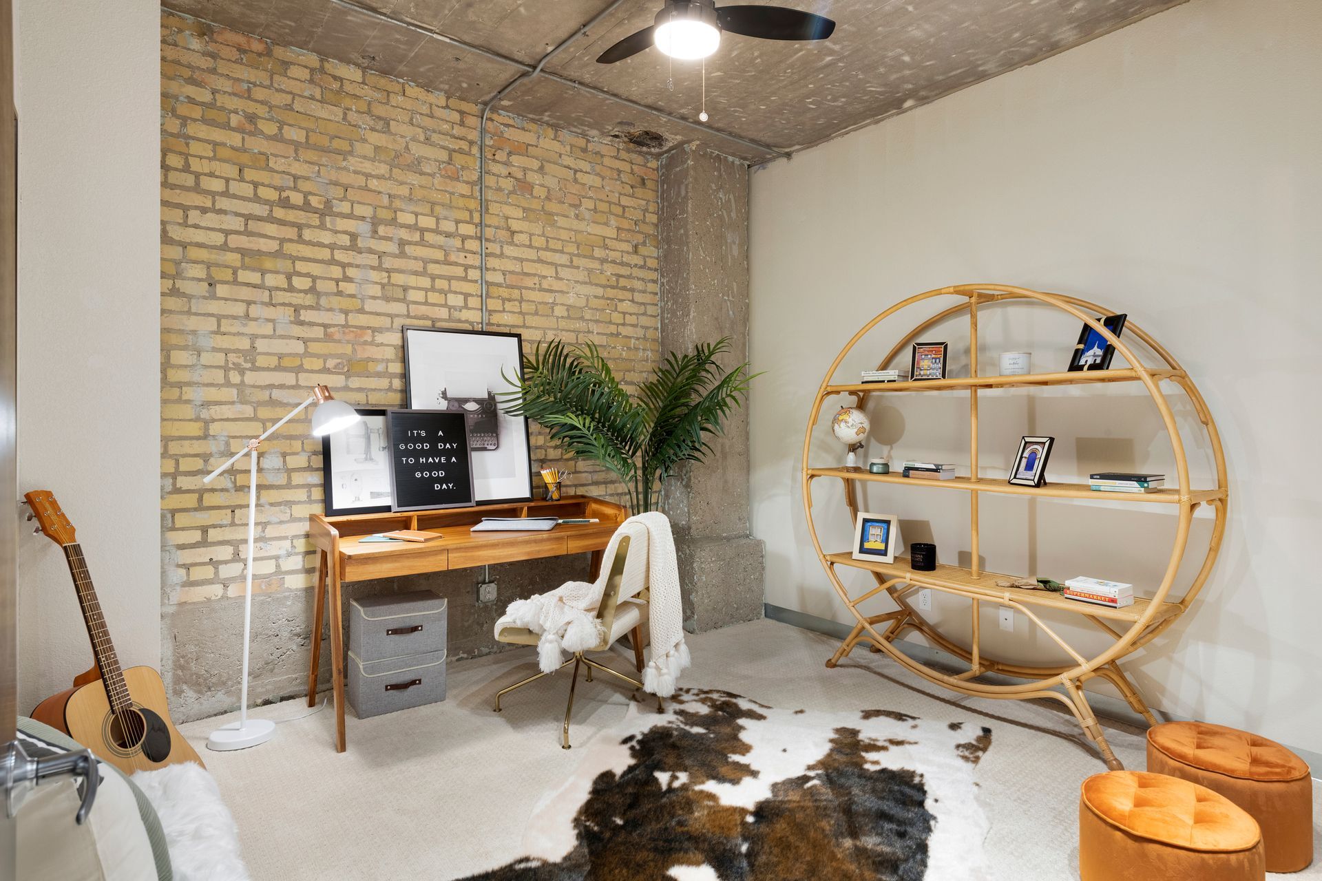 A home office with a wooden desk, circular shelf, patterned rug, and acoustic guitar against a brick accent wall.