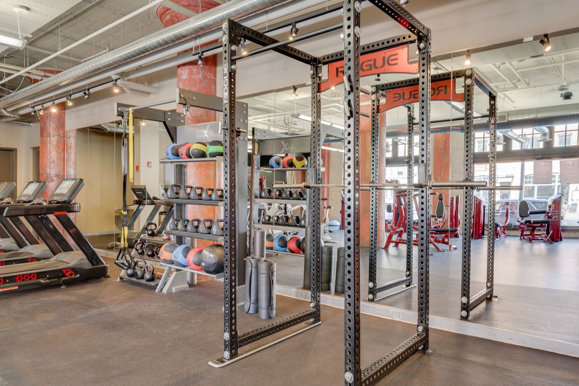 A modern gym featuring several Rogue power racks, rows of kettlebells, and treadmills on a rubber floor.