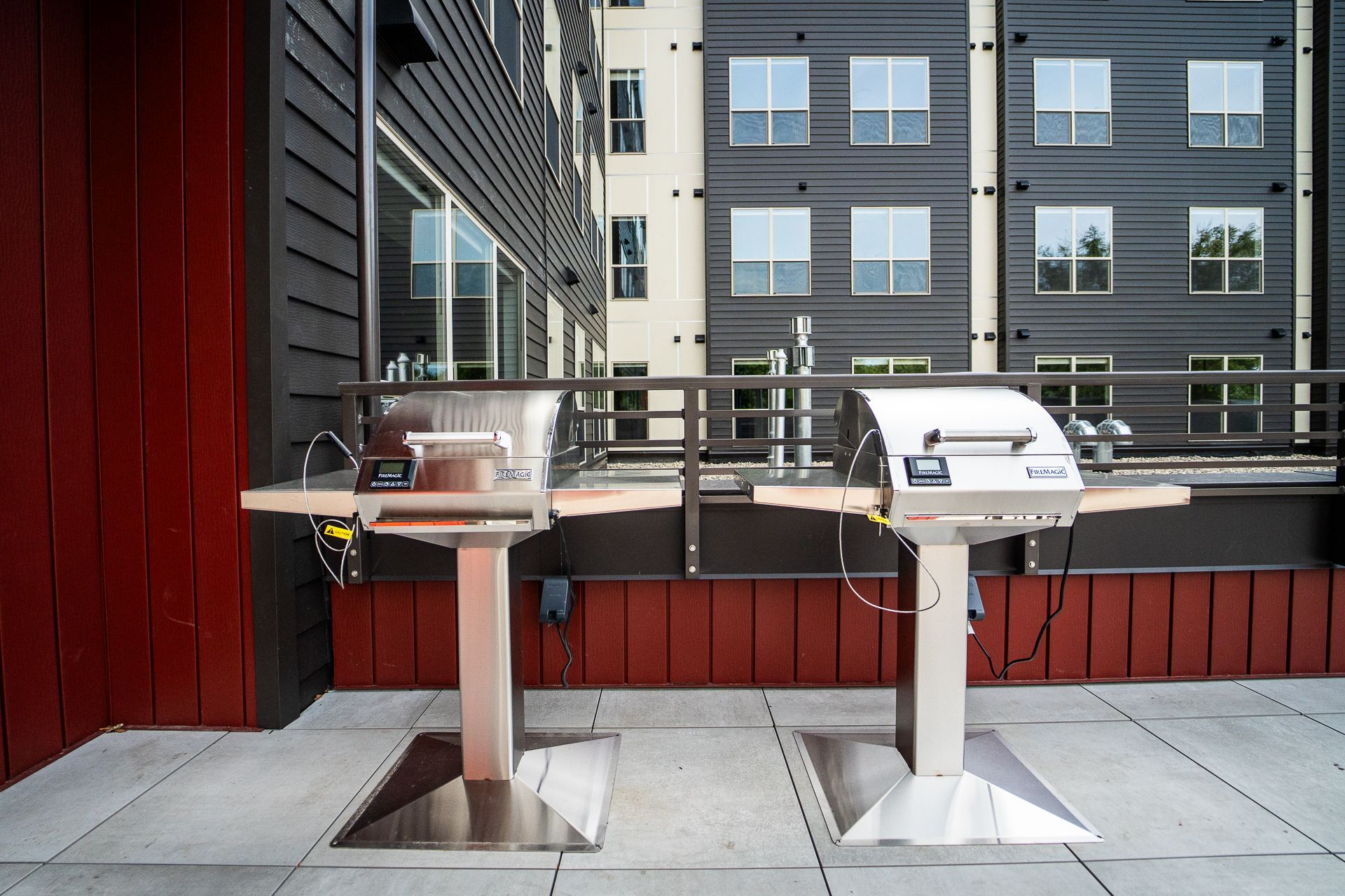 Two stainless steel outdoor grills on a patio terrace in front of a modern apartment building with black and beige siding.