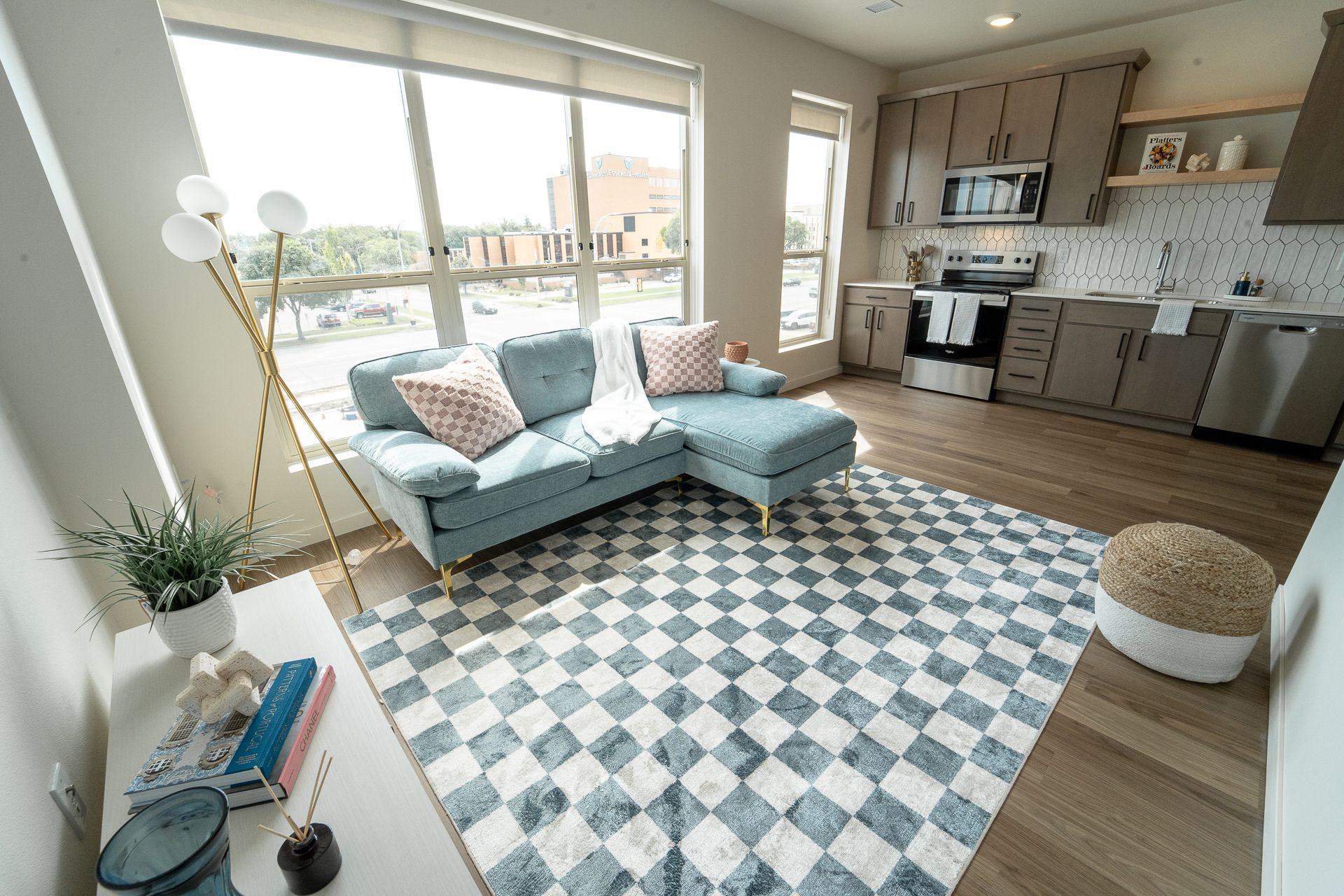 A modern studio apartment with a light blue sectional sofa, a checkered area rug, and an open kitchen with gray cabinets.