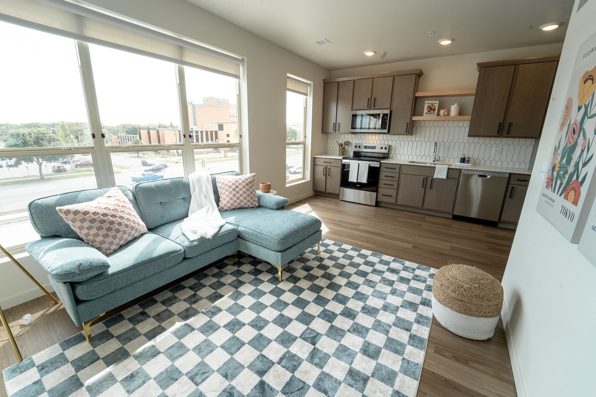 Modern apartment with a light blue sectional sofa, checkered area rug, and an open kitchen with stainless steel appliances.