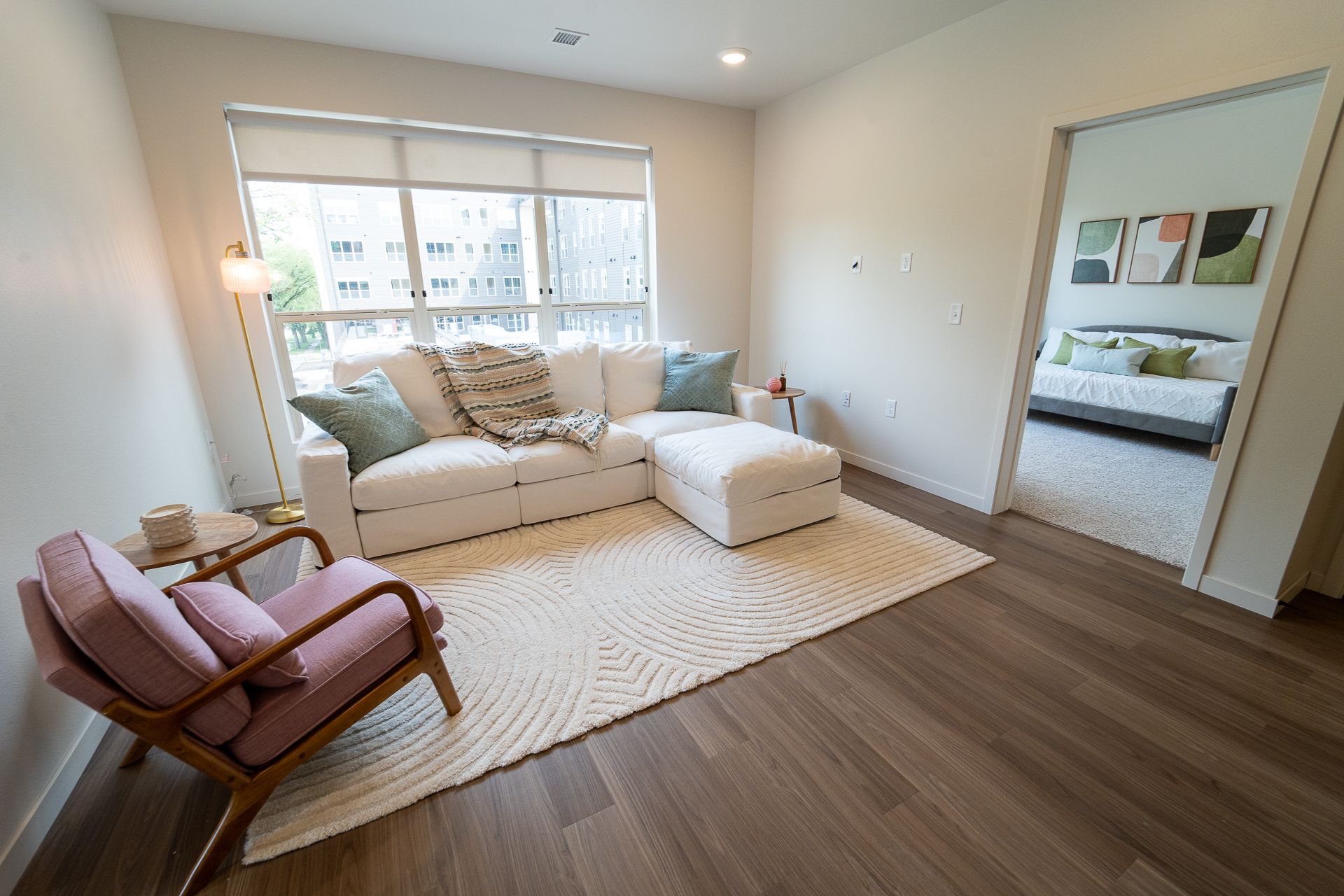 A bright living room with a white sectional sofa, cream rug, pink armchair, and a view into a bedroom with a bed.