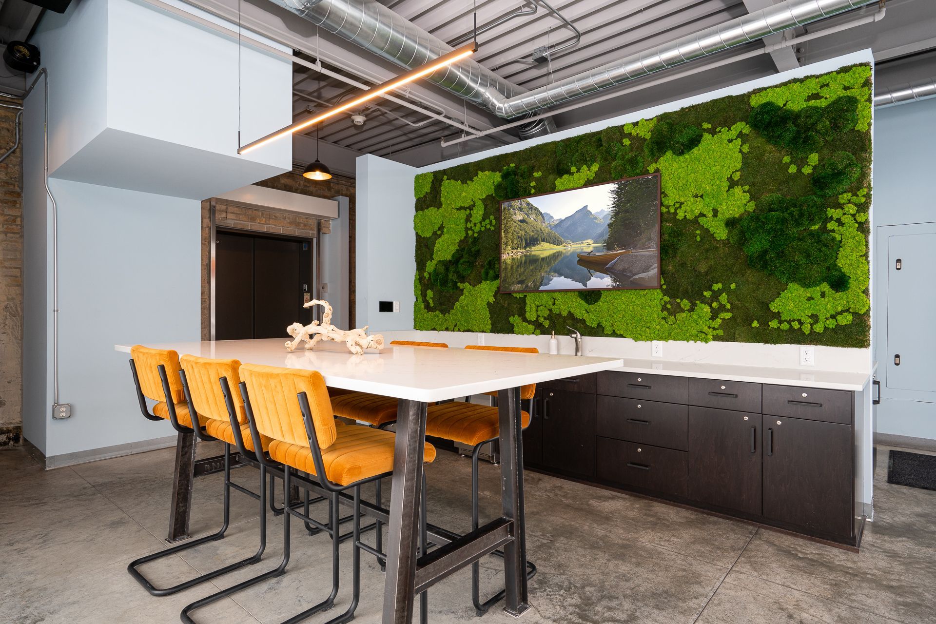 Modern office kitchenette with a high table, yellow stools, and a large wall-mounted moss art piece featuring a landscape.