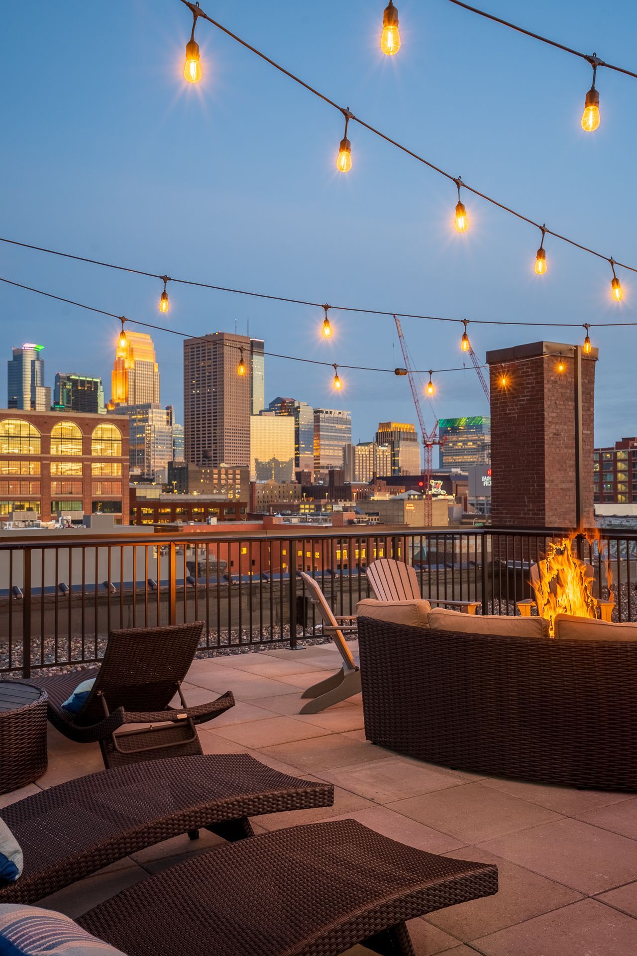 A rooftop terrace with outdoor furniture and a fire pit, overlooking a city skyline at dusk with string lights overhead.