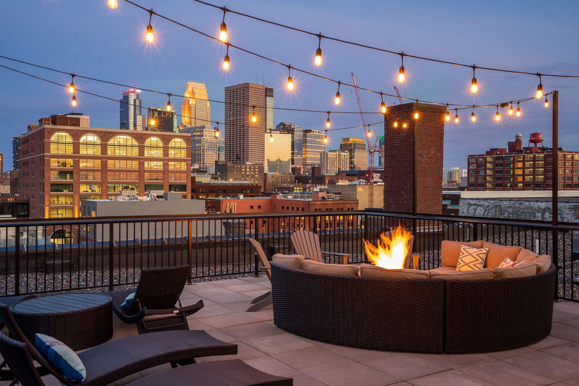 A rooftop lounge at dusk with a circular sofa around a fire pit, string lights, and a view of a city skyline.