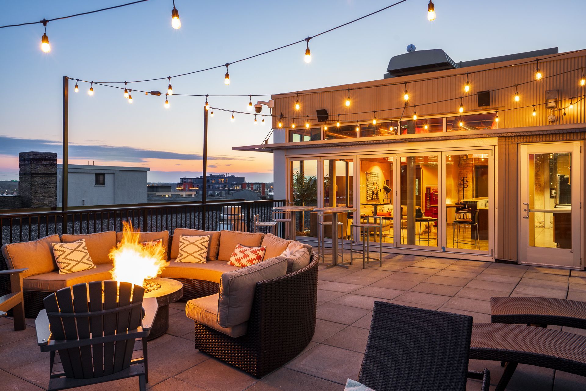 Rooftop patio at dusk with a lit fire pit, outdoor sofa seating, and string lights illuminating the lounge area.