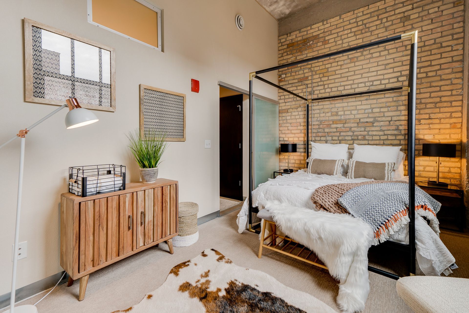 A modern bedroom features a four-poster bed against a brick wall, a wooden sideboard, and a faux-fur rug on the floor.