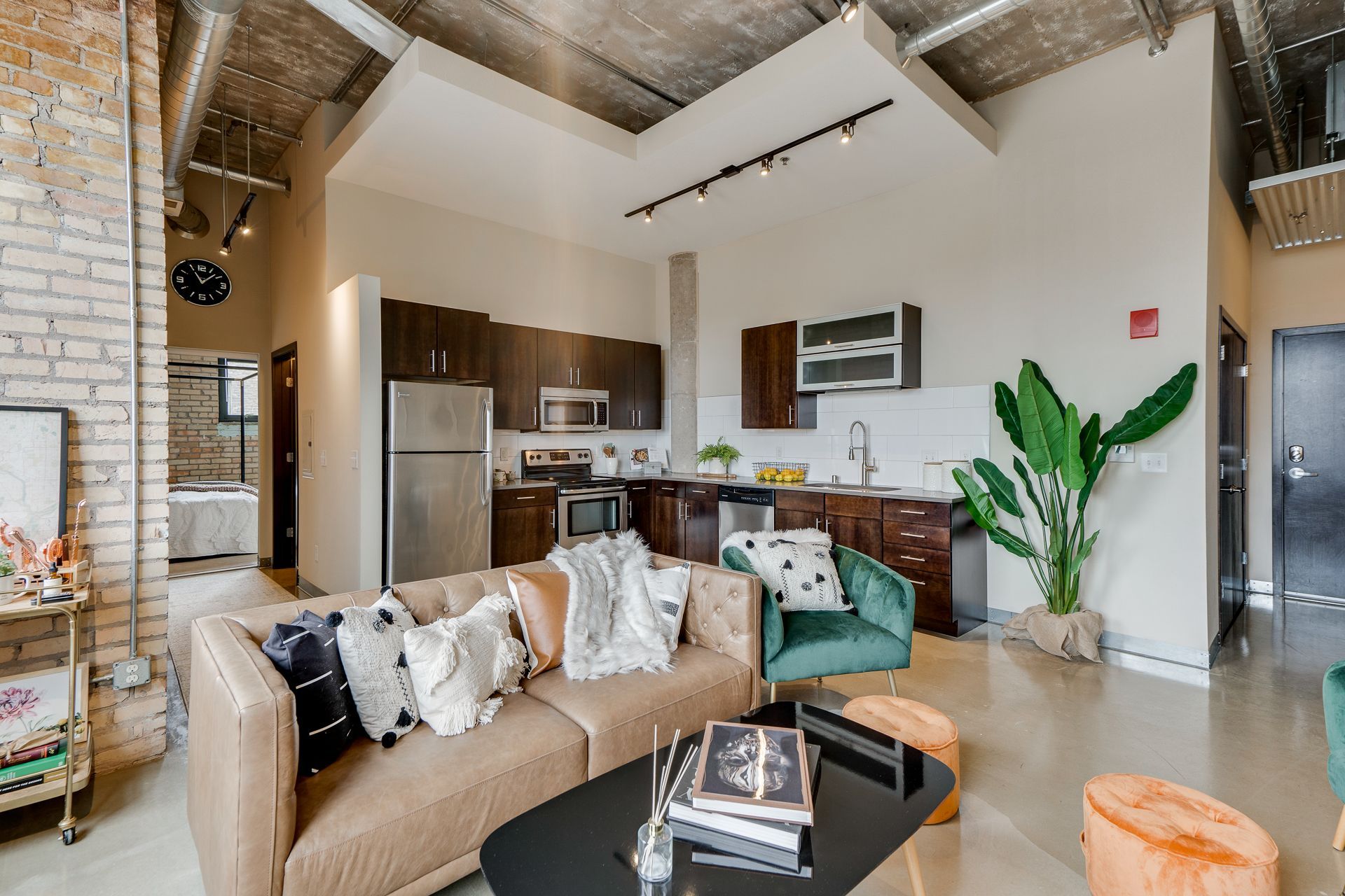 A modern open-plan apartment with a tan sofa, green armchair, and kitchen with dark cabinets and exposed ceiling pipes.