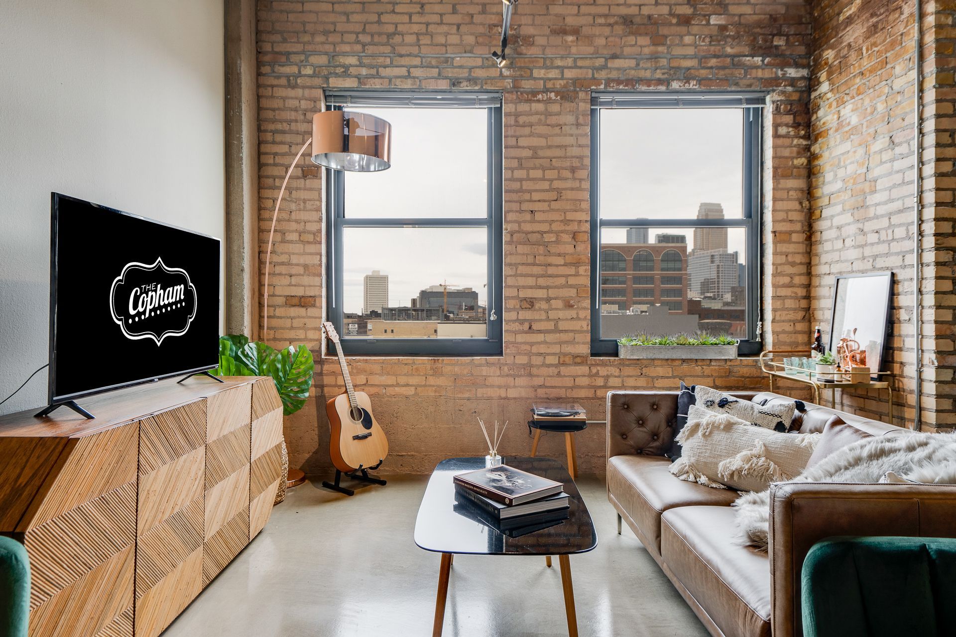 A cozy living room with a brown leather sofa, wooden cabinet, guitar, floor lamp, and city view through brick-walled windows.