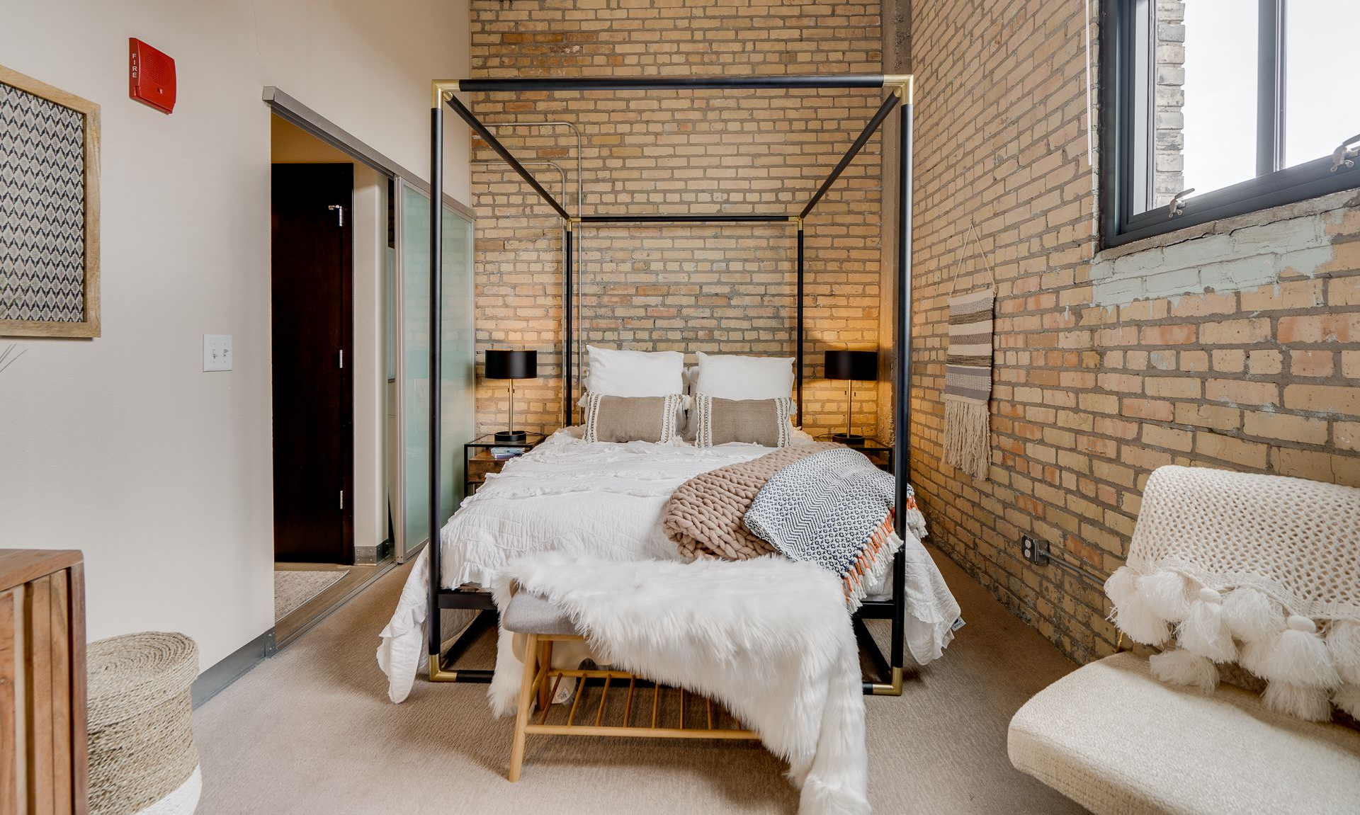 A bedroom with an exposed brick wall, a black four-poster bed with white bedding, and a cozy chair in the foreground.